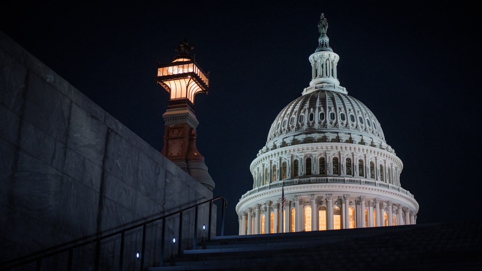 The illuminated dome of the U.S. Capitol at night, seen from a stone staircase with a glowing lantern-style lamp post at left.