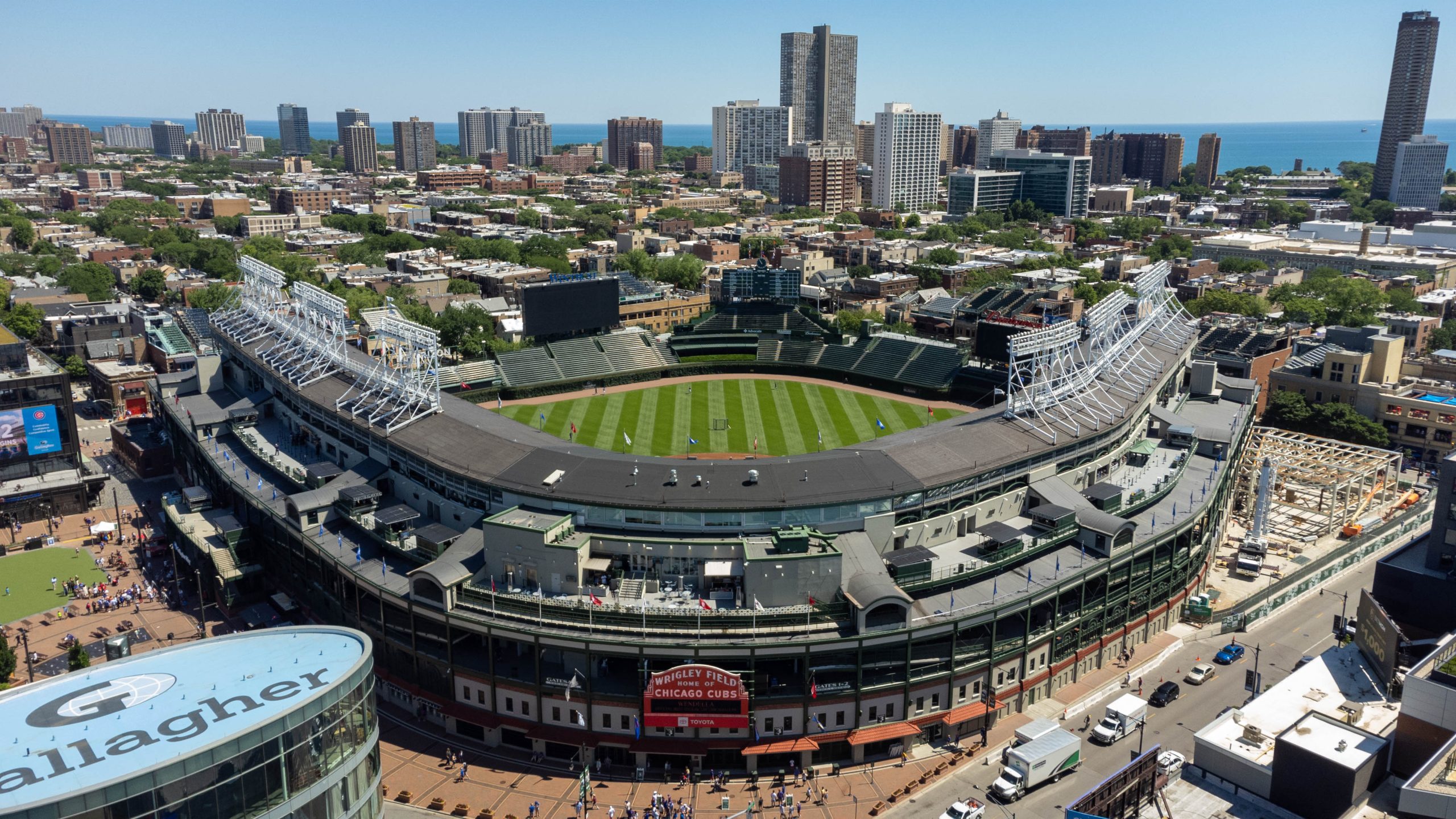 Aerial view of Wrigley Field in Chicago on a sunny day, with the empty green field, the red entrance marquee and the North Side neighborhood and Lake Michigan beyond.