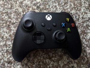 A black Xbox game controller with a lit Xbox logo button and colored A, B, X and Y buttons, lying on a speckled granite surface.