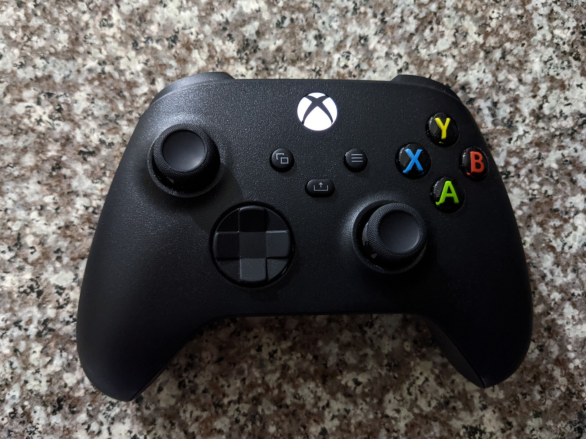 A black Xbox game controller with a lit Xbox logo button and colored A, B, X and Y buttons, lying on a speckled granite surface.