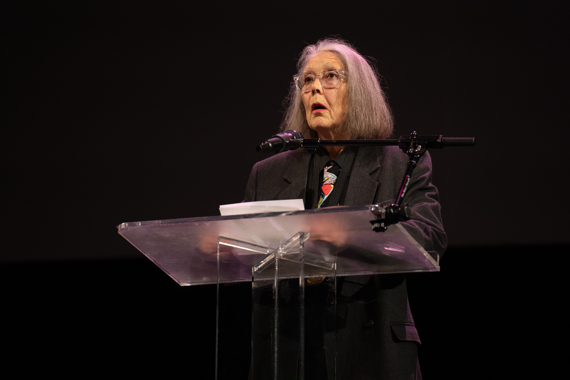 Anne Carson, a woman with long gray hair and glasses, speaking at a clear lectern with a microphone against a dark background.