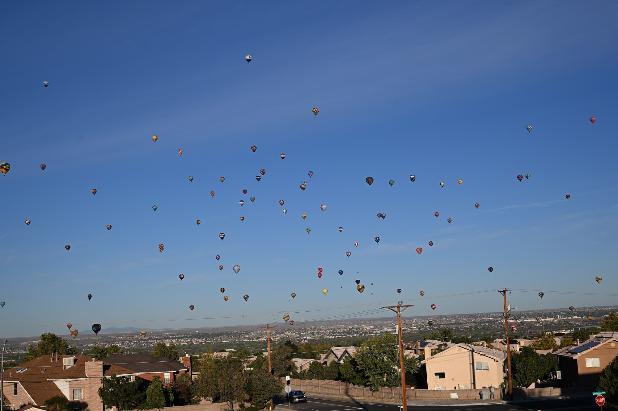 Dozens of hot air balloons in a blue morning sky above houses and utility poles on the edge of Albuquerque, with the city spread out below.