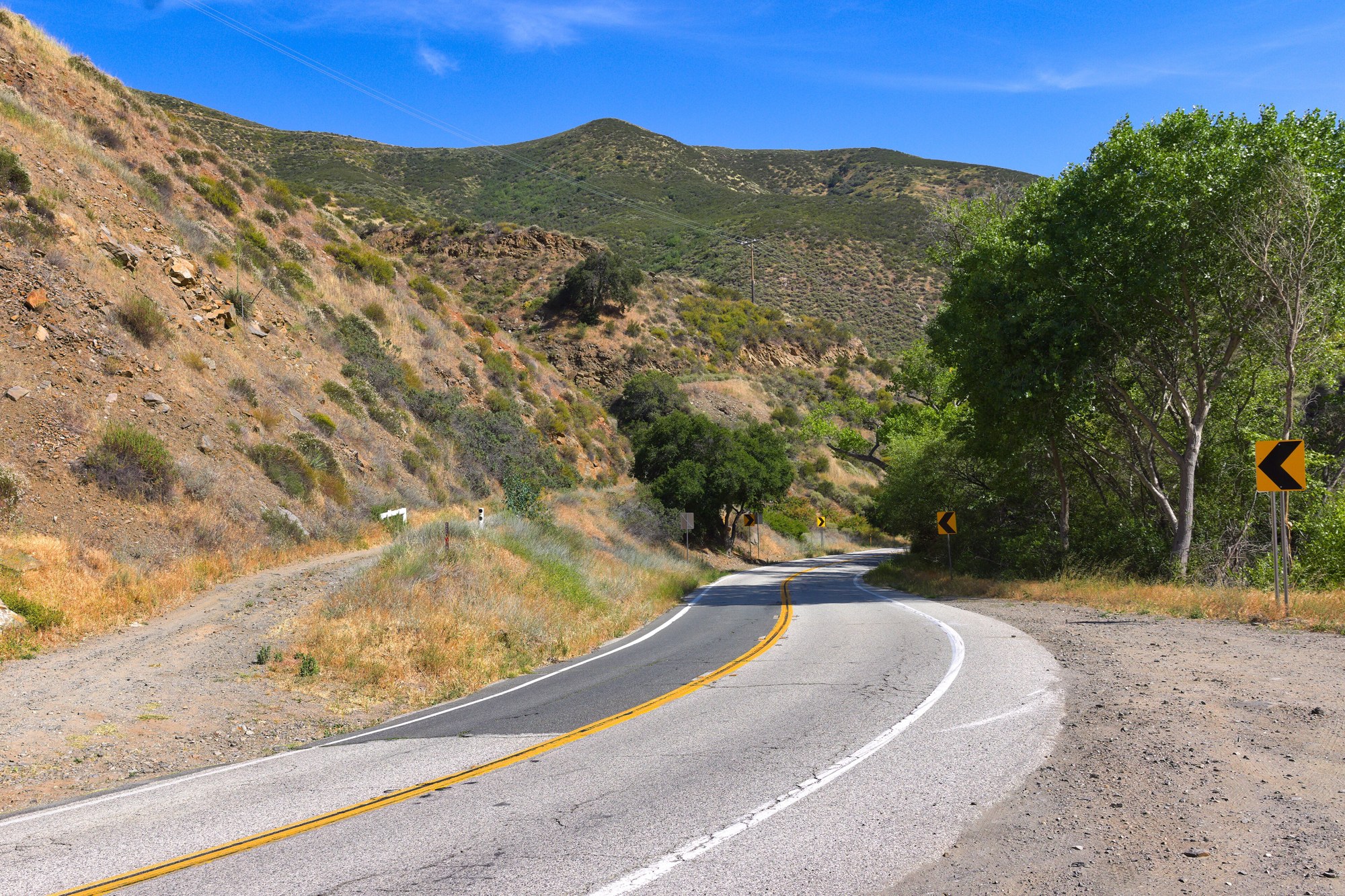 A two-lane mountain road with a double yellow line curving through dry, brush-covered hills under a blue sky, with green trees along one side.