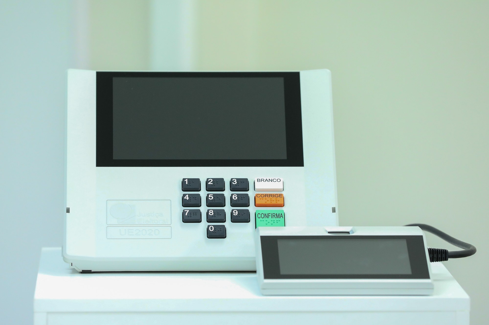 A Brazilian electronic voting machine, model UE2020, with a numeric keypad and BRANCO, CORRIGE and CONFIRMA buttons, beside the voter-identification terminal.