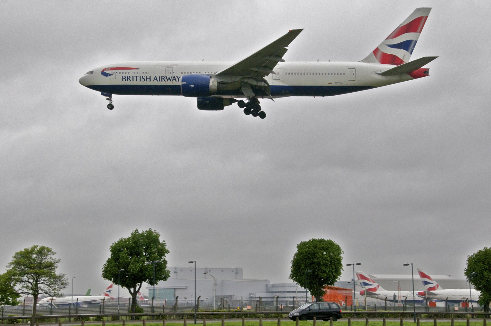 A British Airways Boeing 777 with its landing gear down approaches a runway under a gray sky, with other British Airways aircraft parked in the background.
