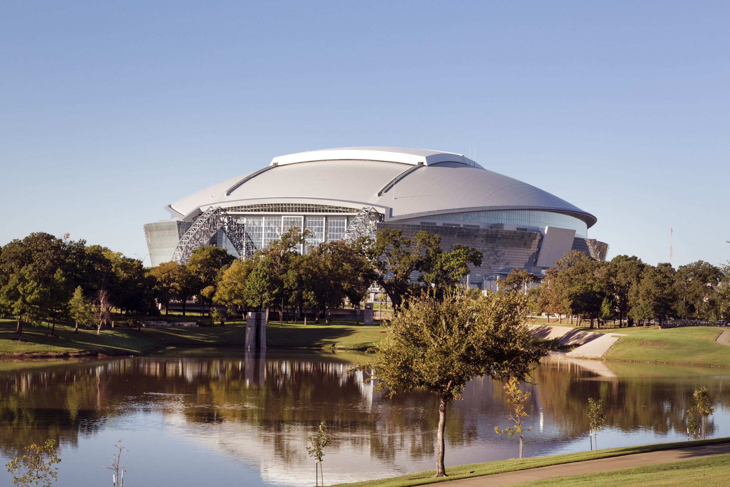 A large domed stadium with a retractable roof seen across a pond and trees under a clear blue sky.