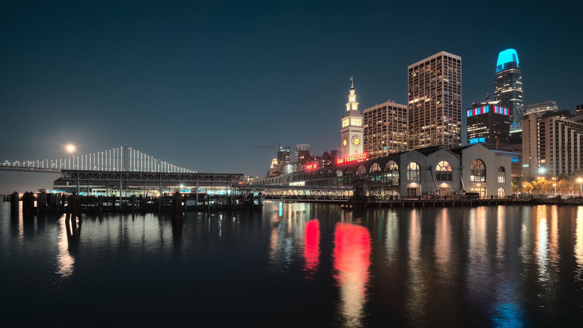 The lit clock tower of the San Francisco Ferry Building and downtown towers at night, reflected in dark water beside the Bay Bridge.