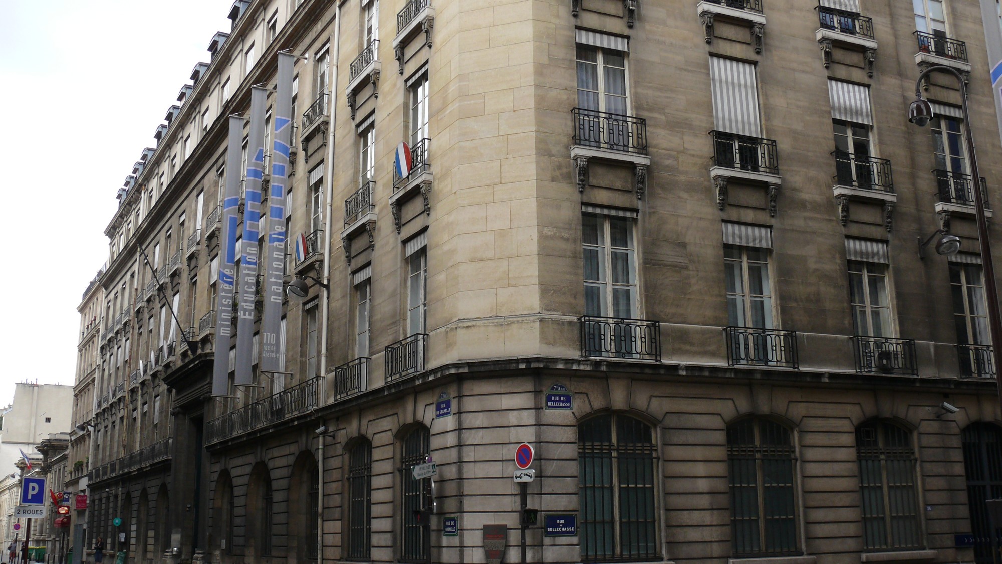 A corner of a cream-colored stone building in Paris with tall windows, iron balconies and vertical banners reading Ministère de l'Éducation nationale, 110.