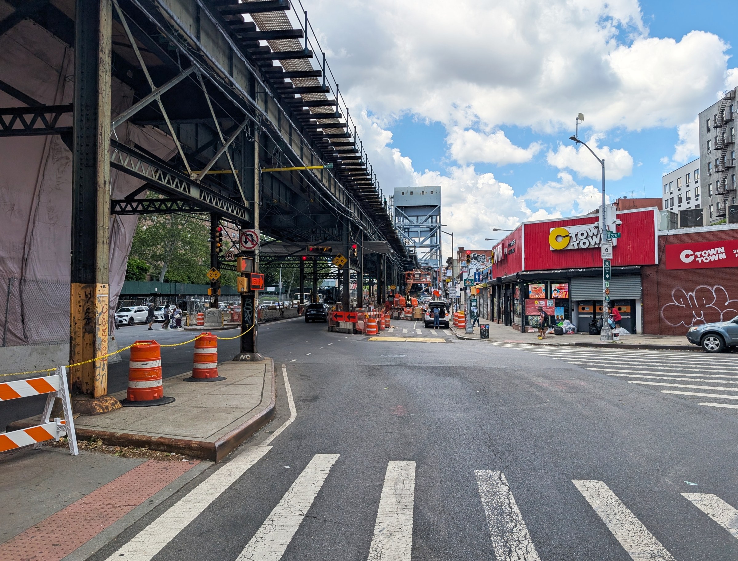 Broadway in Marble Hill under an elevated subway structure, with a crosswalk, orange construction barrels and a red storefront on a sunny day.