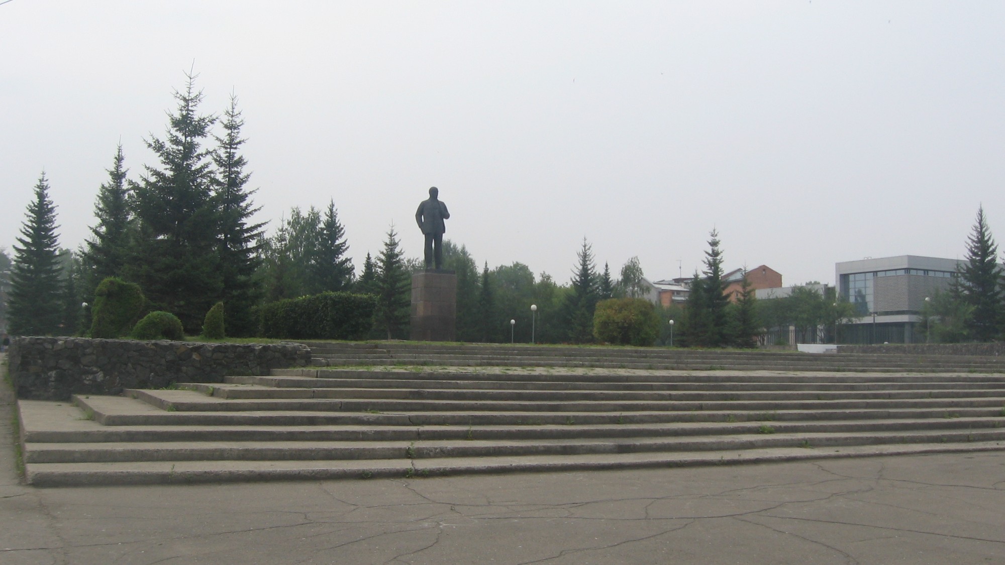 A bronze statue of a standing figure on a stone pedestal above wide concrete steps in a town square, with spruce trees and low buildings under a hazy sky.