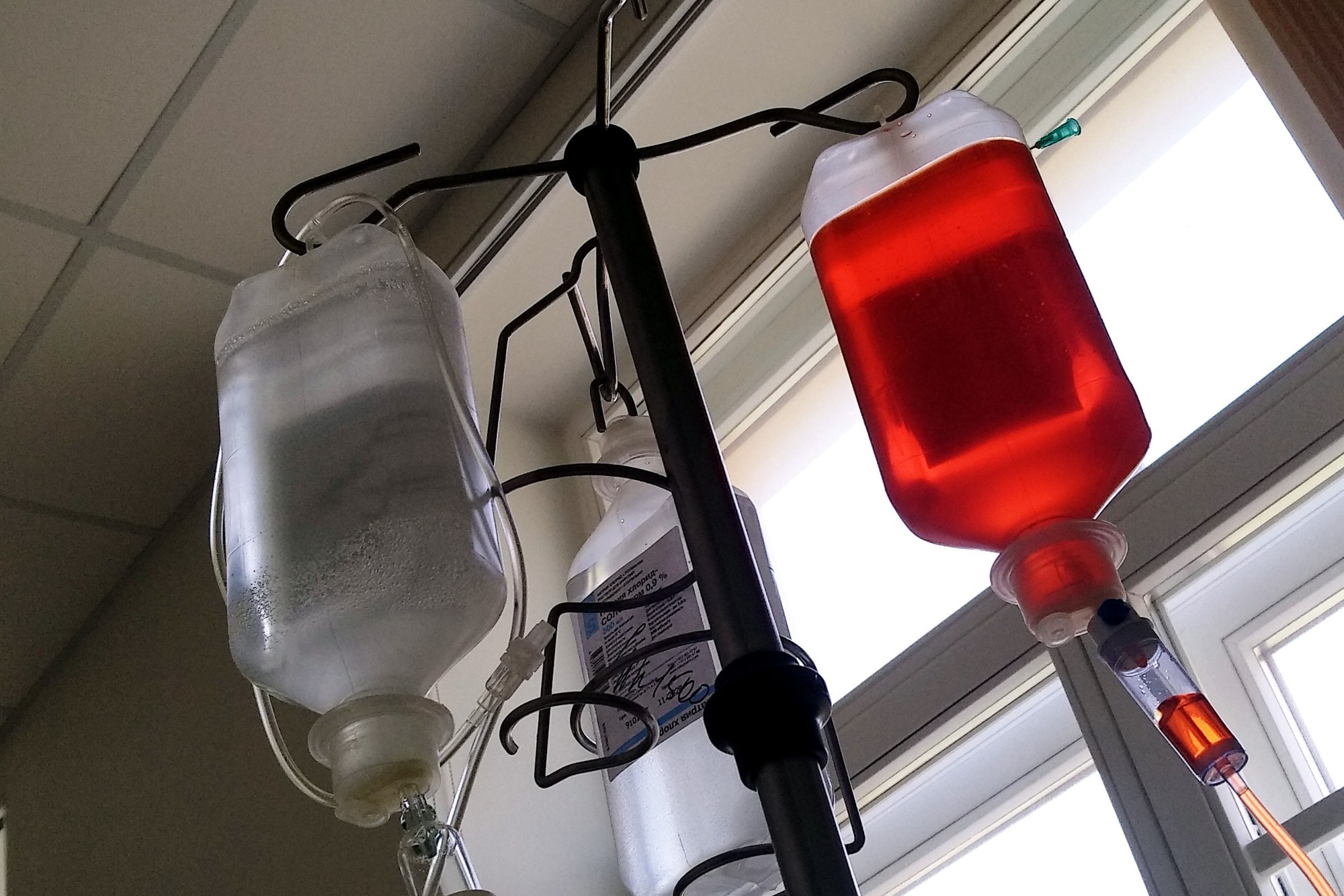 Intravenous infusion bags, one deep red, hanging from a hospital IV pole beside a window.