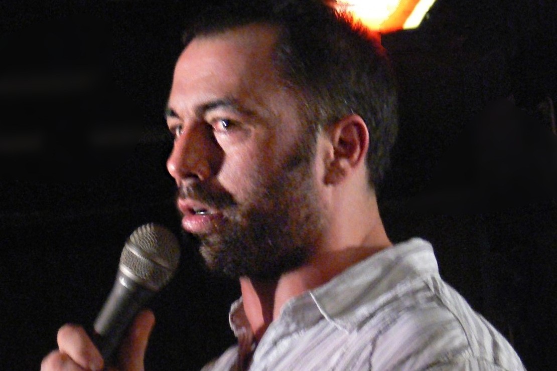 A bearded man in a striped shirt speaking into a handheld microphone on a dark stage.