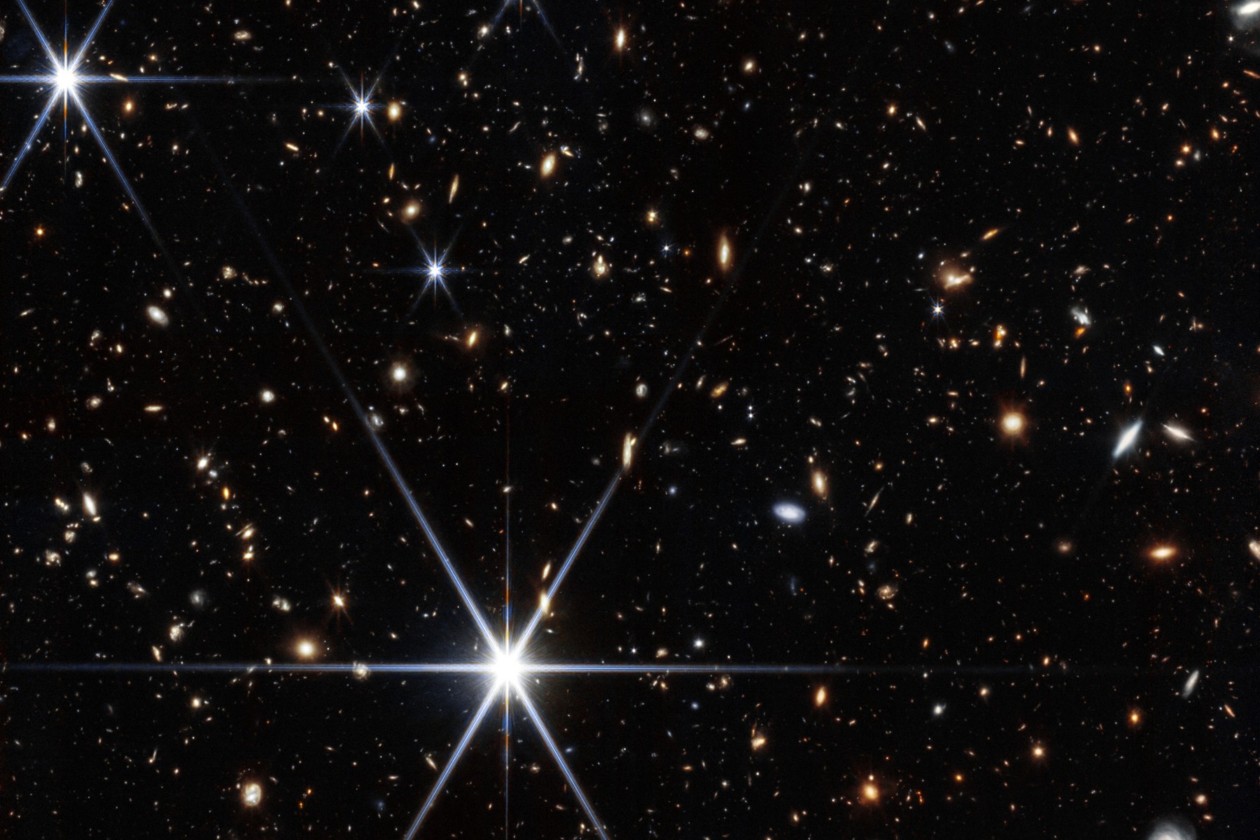 A dense field of small galaxies of many shapes and colors on black space, with bright blue-white stars showing long diffraction spikes, in a James Webb Space Telescope near-infrared image.
