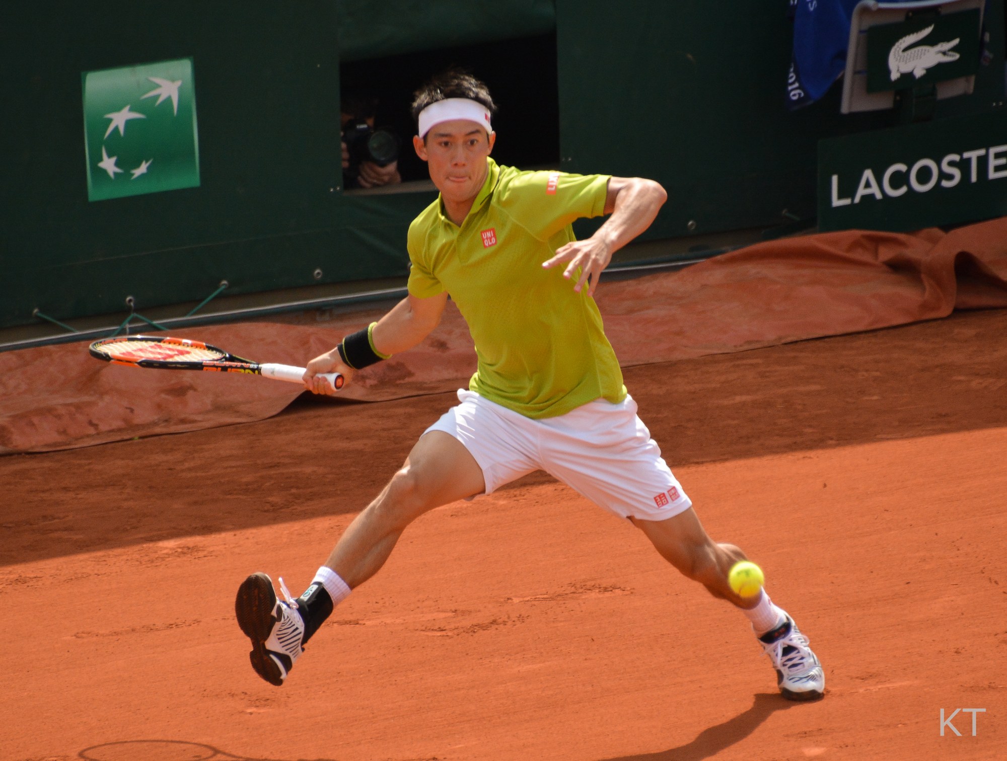 Kei Nishikori stretches wide to hit a forehand on clay at the 2016 French Open, wearing a yellow-green shirt and a white headband.