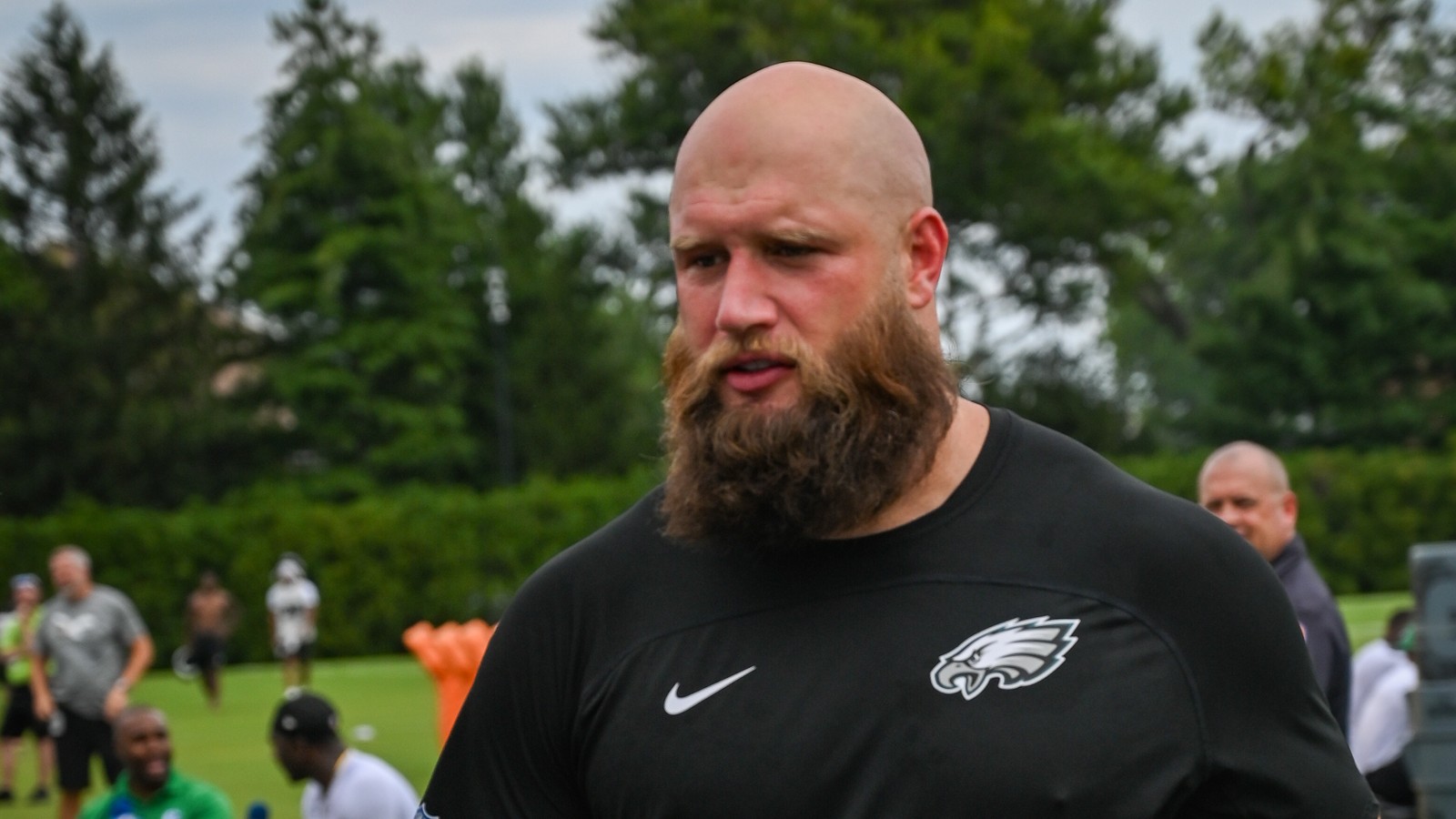 Eagles offensive lineman Lane Johnson, a bald, bearded player in a black Eagles shirt and shorts, walks across a grass practice field during training camp.