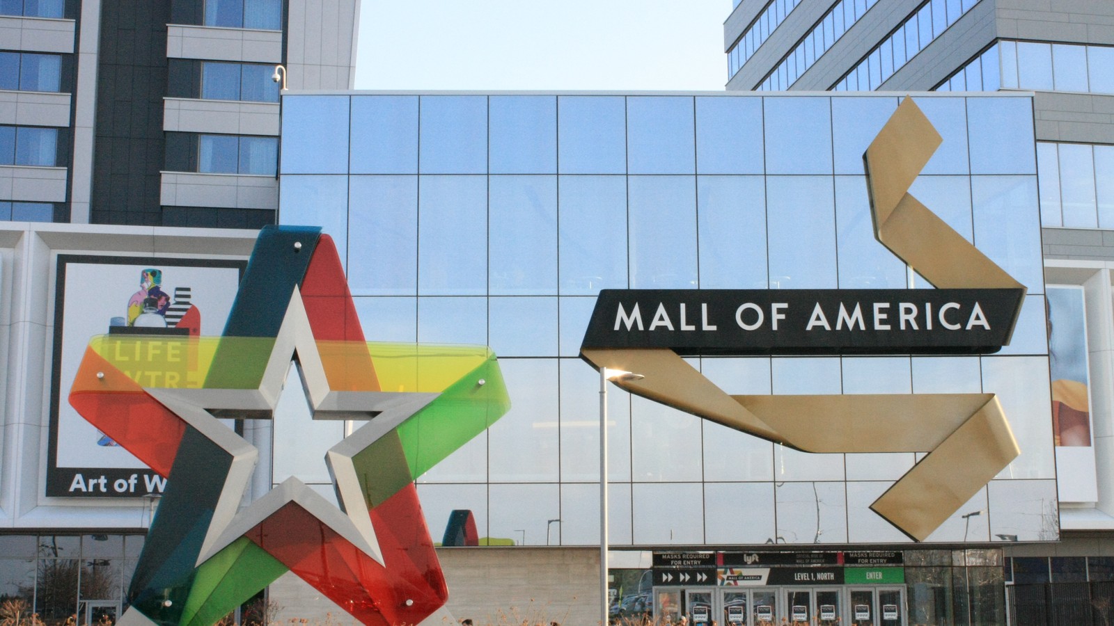 The glass front entrance of the Mall of America in Bloomington, Minnesota, with the mall's name on a black sign beside a gold ribbon sculpture and a colorful glass star sculpture.