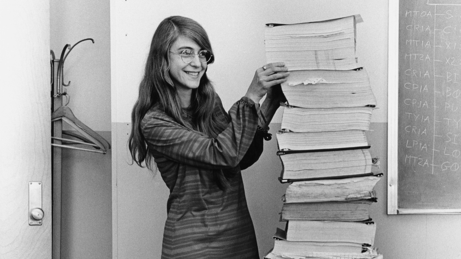 Margaret Hamilton in 1969, a young woman with long hair and glasses smiling as she stands beside a tall stack of printed listings of the Apollo software her MIT team wrote.