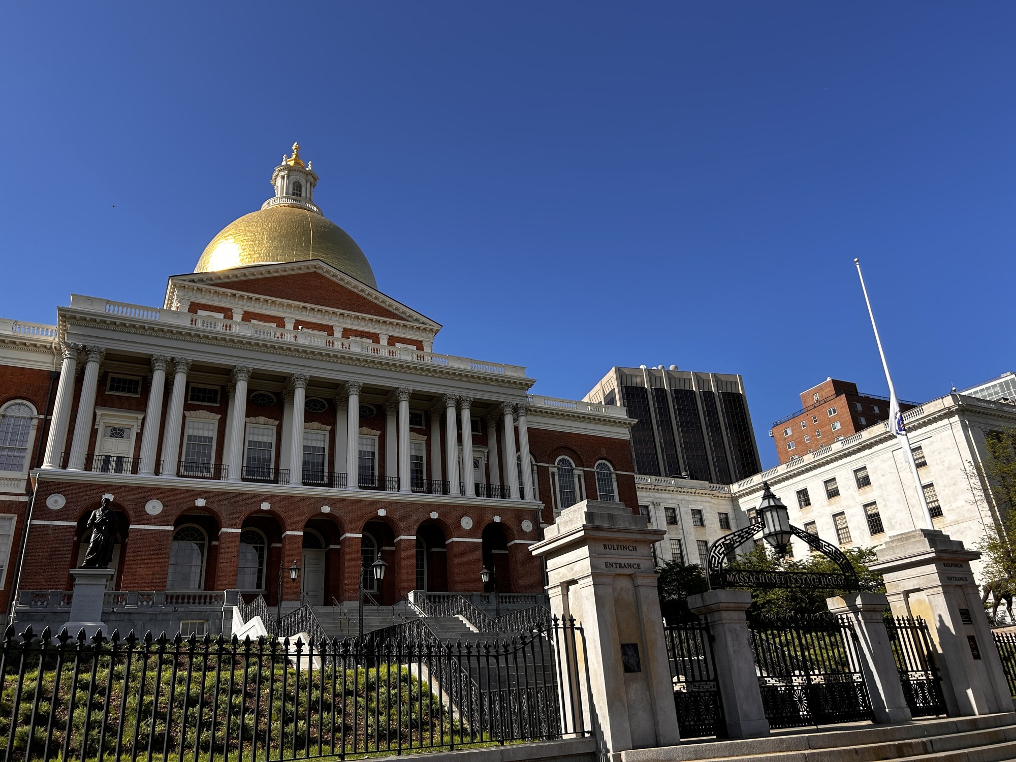The Massachusetts State House in Boston, a red-brick building with white columns and a gold dome, seen beyond an iron fence and its Bulfinch entrance gate under a clear blue sky.