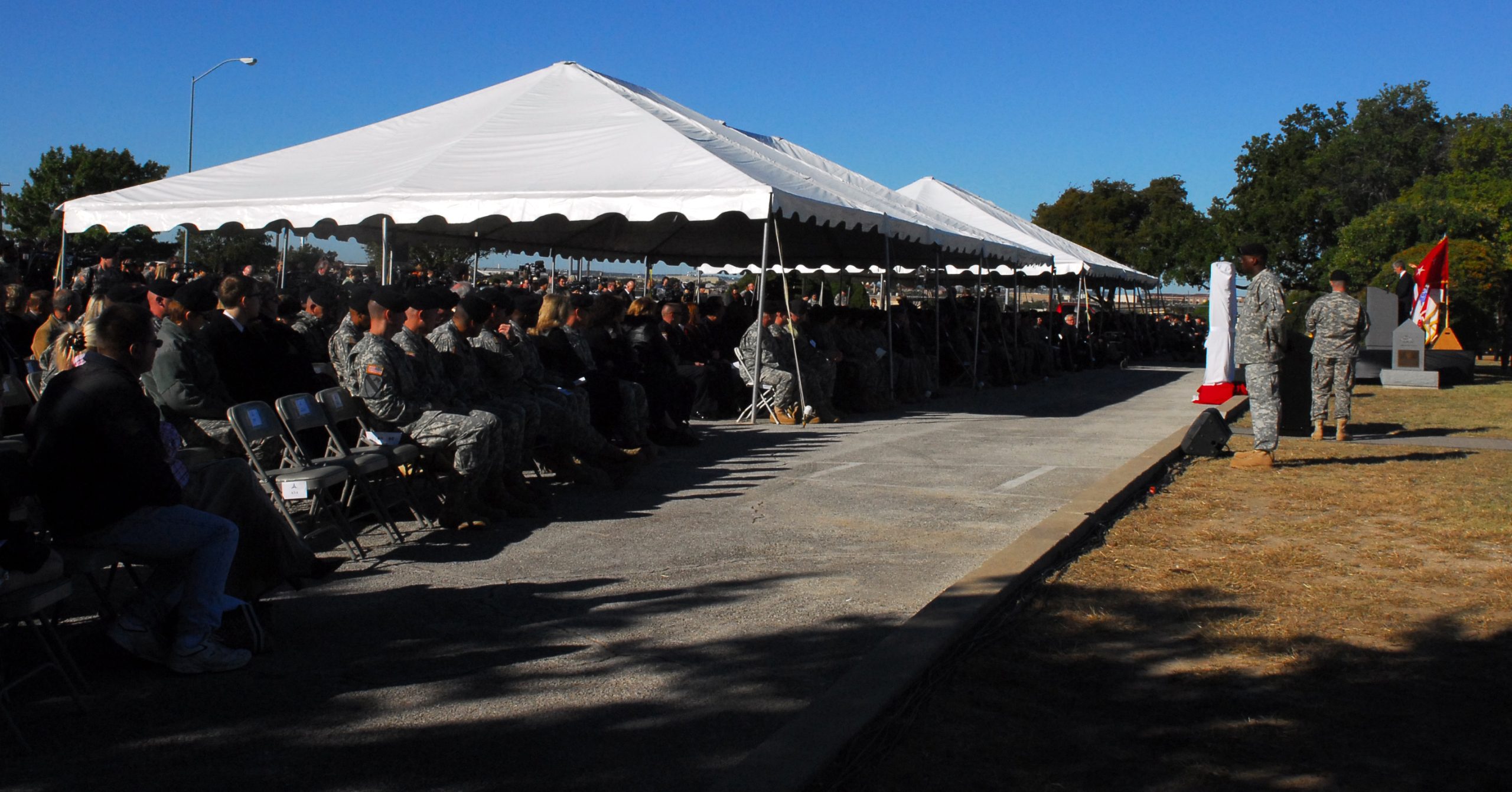 Rows of soldiers and families seated under white tents at an outdoor memorial ceremony at Fort Hood, Texas, with a draped monument and flags at the right under a blue sky.