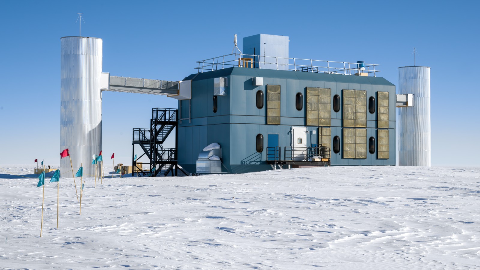 The IceCube Laboratory at the South Pole: a blue-grey two-storey building with a railed roof, flanked by two tall metal cylinders, on flat snow under a clear blue sky, with small colored flags on stakes in the foreground.