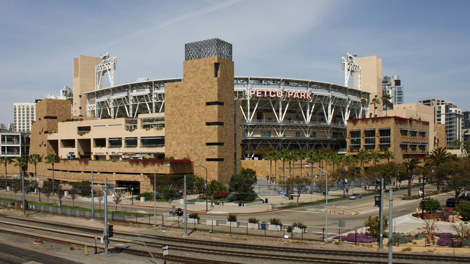 The exterior of Petco Park in San Diego, the Padres' ballpark, seen across the railway and trolley tracks, with the PETCO PARK sign on the stadium's steel frame and sandstone-colored towers under a pale blue sky.