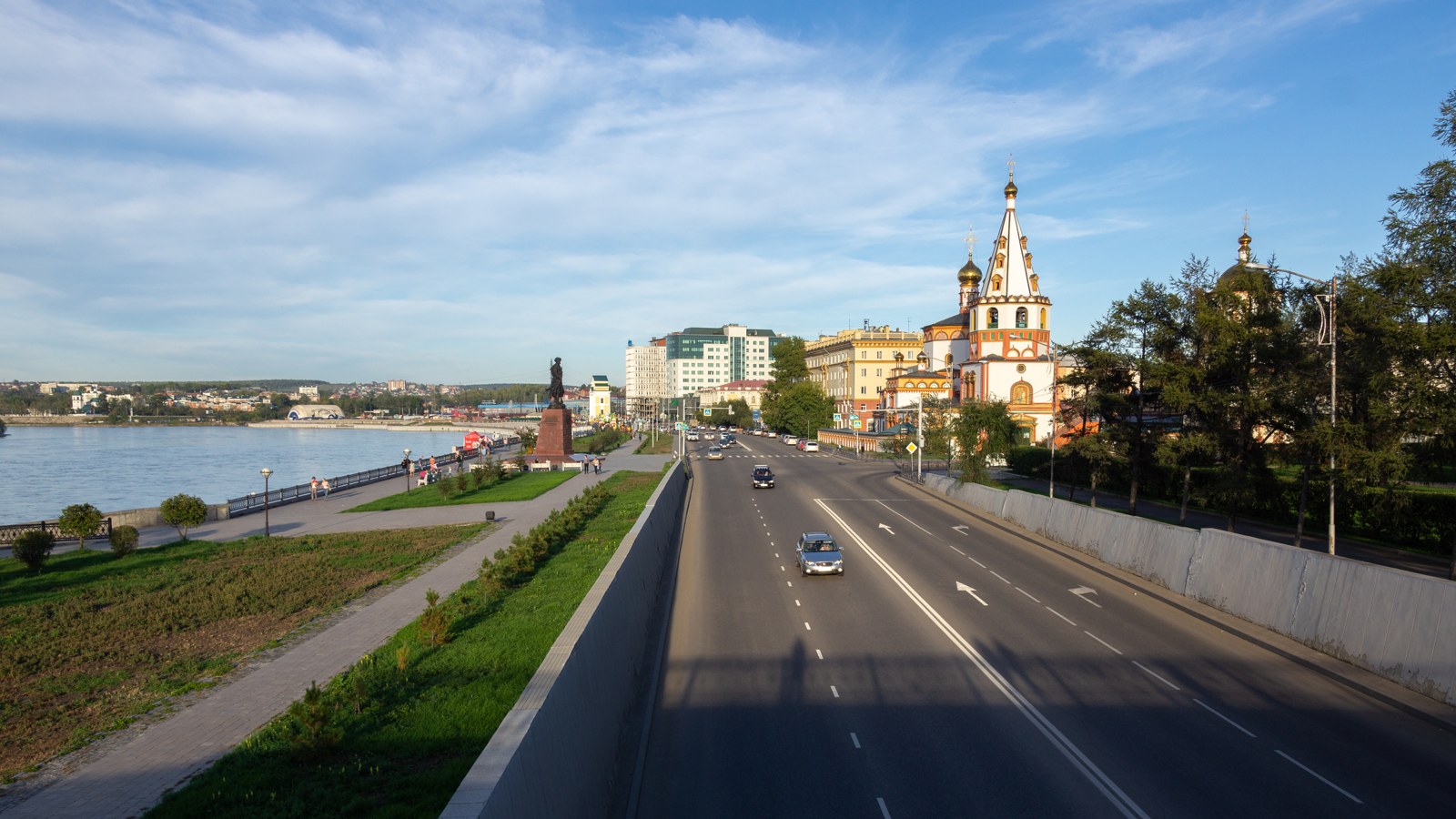 A riverside embankment and a multi-lane road in a Siberian city, with a statue near the water and a white-and-gold domed church on the right under a blue sky.
