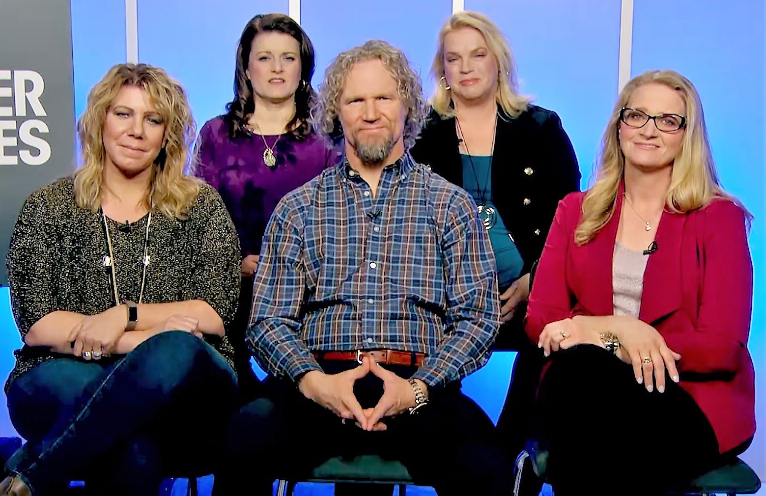 A man in a plaid shirt seated in the center of a television studio set with four women around him, two seated in front and two standing behind.