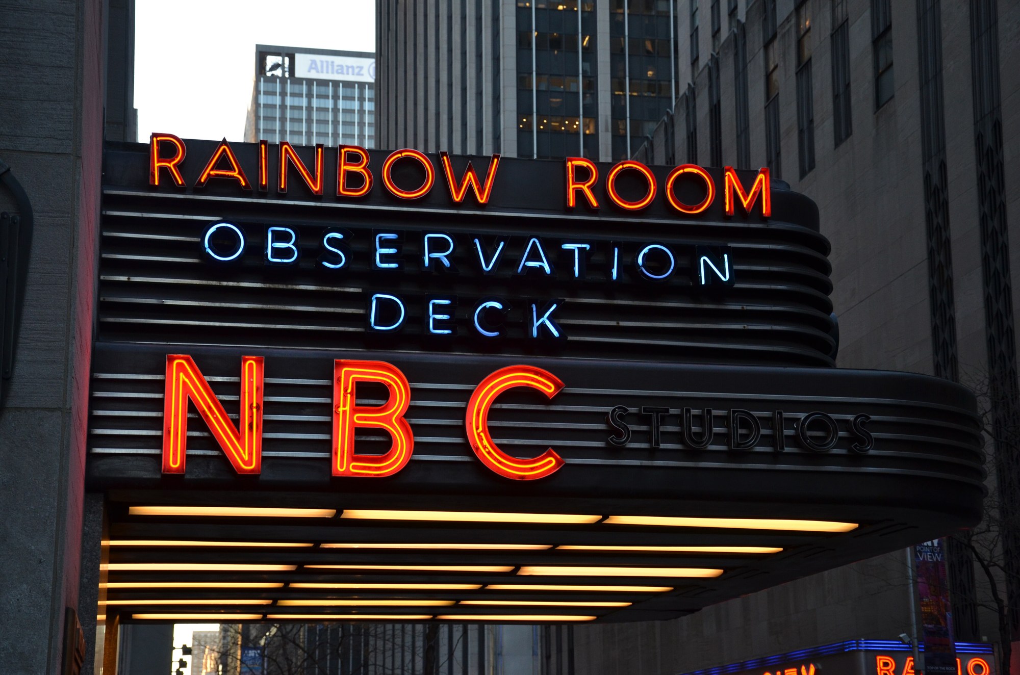 The red neon NBC Studios sign on the curved marquee at Rockefeller Center in New York, with Rainbow Room and Observation Deck signs above it.