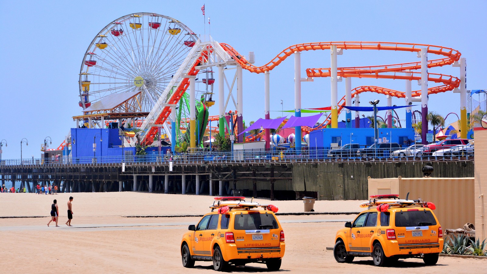 The Santa Monica Pier amusement park with its Ferris wheel and orange roller coaster, seen from a sandy beach where two people walk past two yellow lifeguard vehicles under a clear sky.