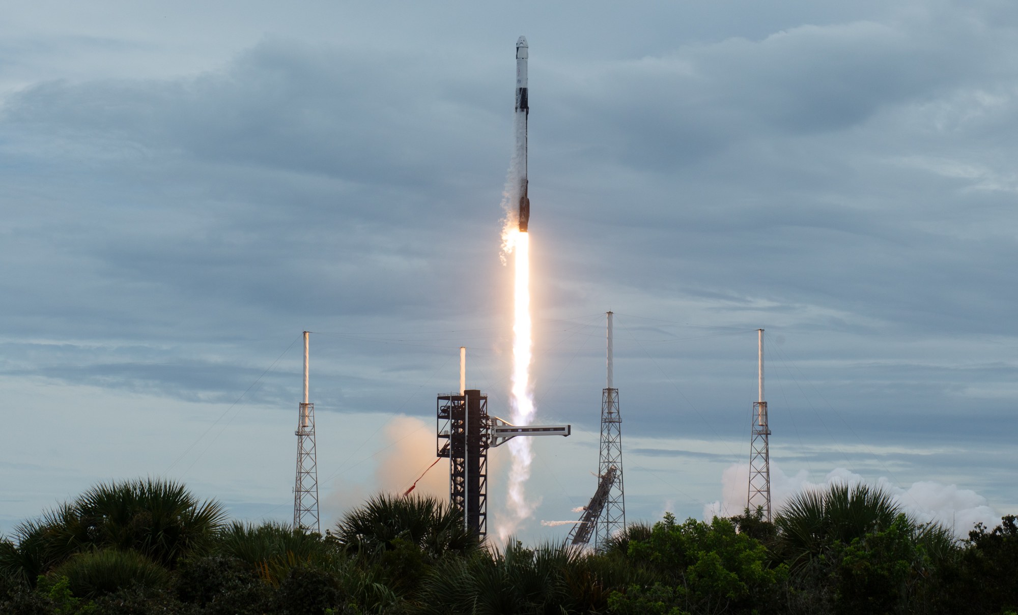A SpaceX Falcon 9 rocket rises on a column of flame from Space Launch Complex 40 at Cape Canaveral under a cloudy sky, with launch towers and palm trees in the foreground.