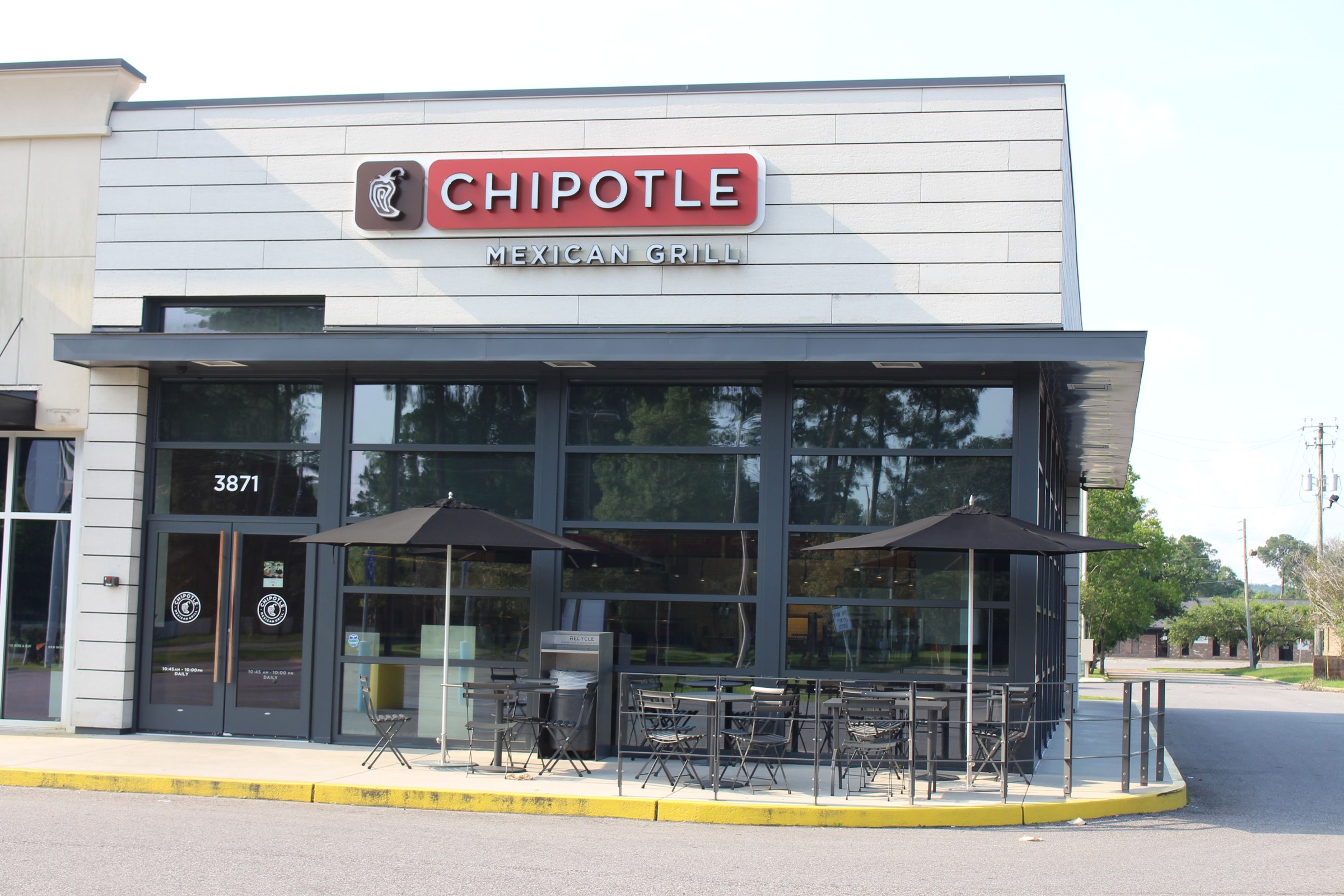 The front of a Chipotle Mexican Grill restaurant with a red Chipotle sign, large windows and outdoor tables with umbrellas.