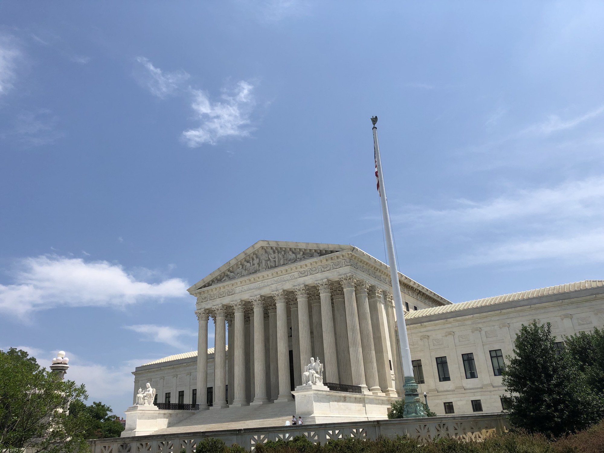 The Supreme Court building in Washington, D.C., seen from the side with its columns, pediment and a flagpole under a blue sky.