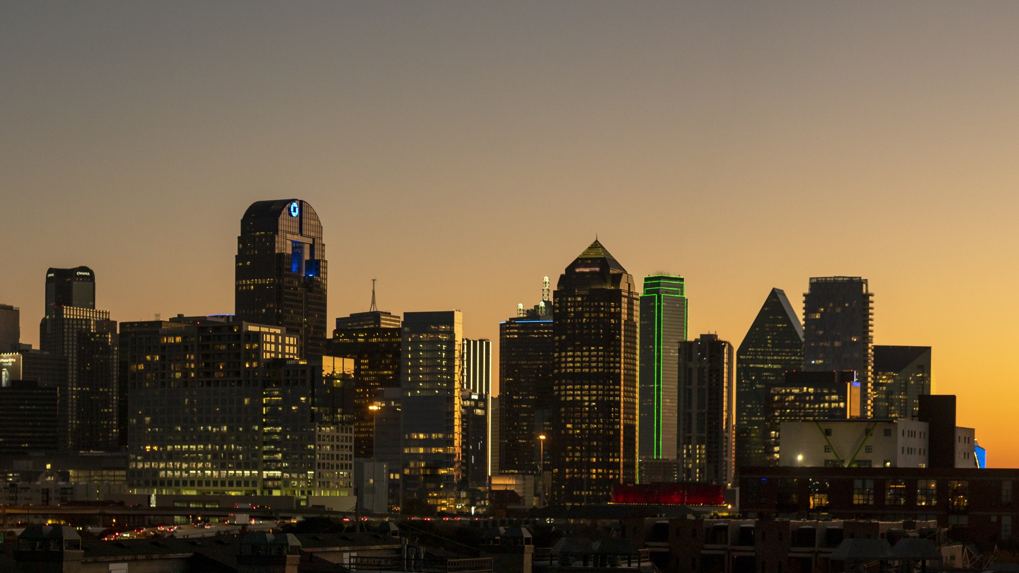 The downtown Dallas skyline at dusk, lit towers silhouetted against an orange and gray sky.