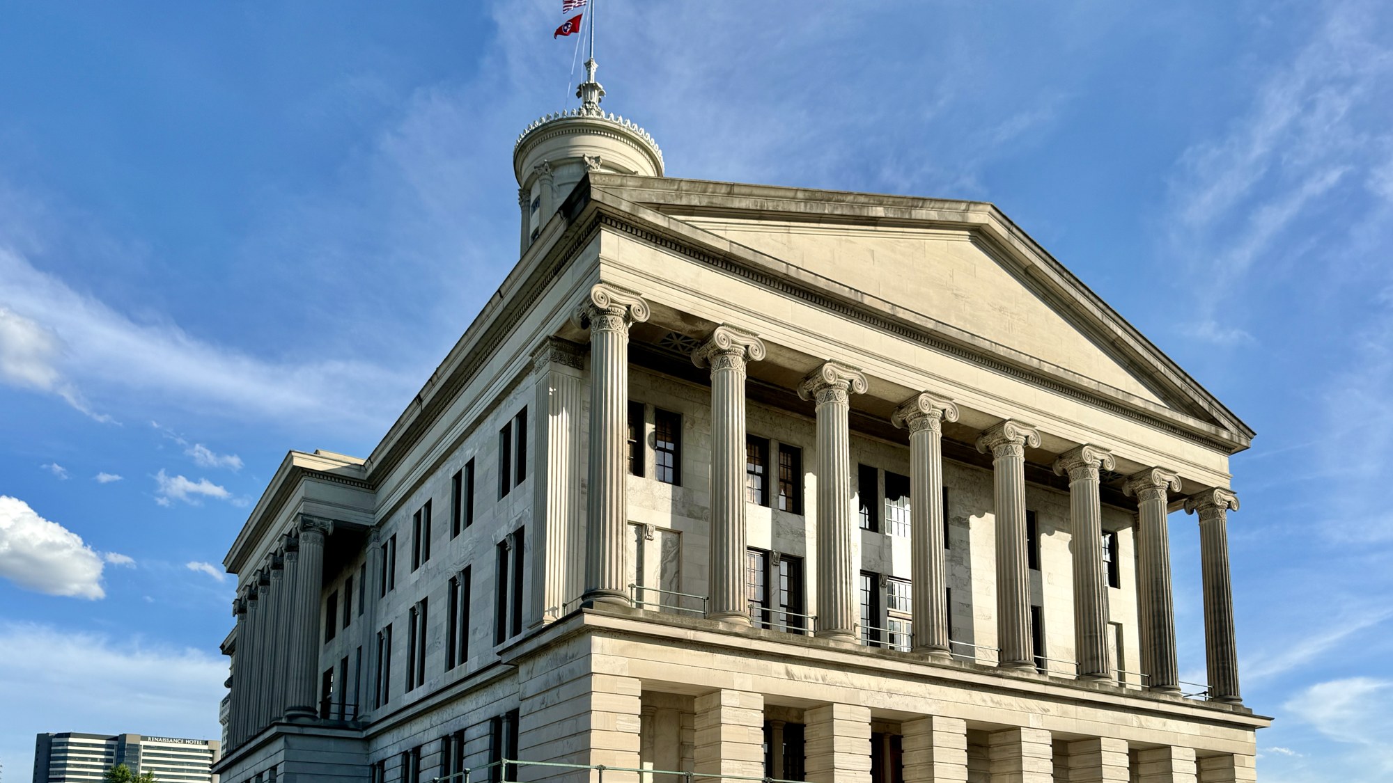 The Greek Revival Tennessee State Capitol in Nashville, seen from a corner under a blue sky with the state flag flying.