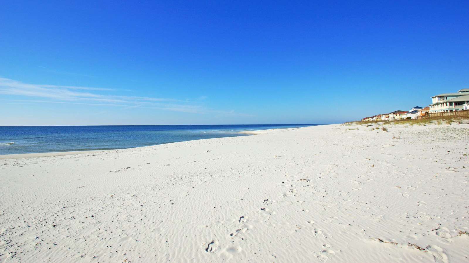 A wide, empty white-sand beach on the Gulf of Mexico at Gulf Shores, Alabama, with calm blue water, a clear sky and beachfront houses behind the dunes.