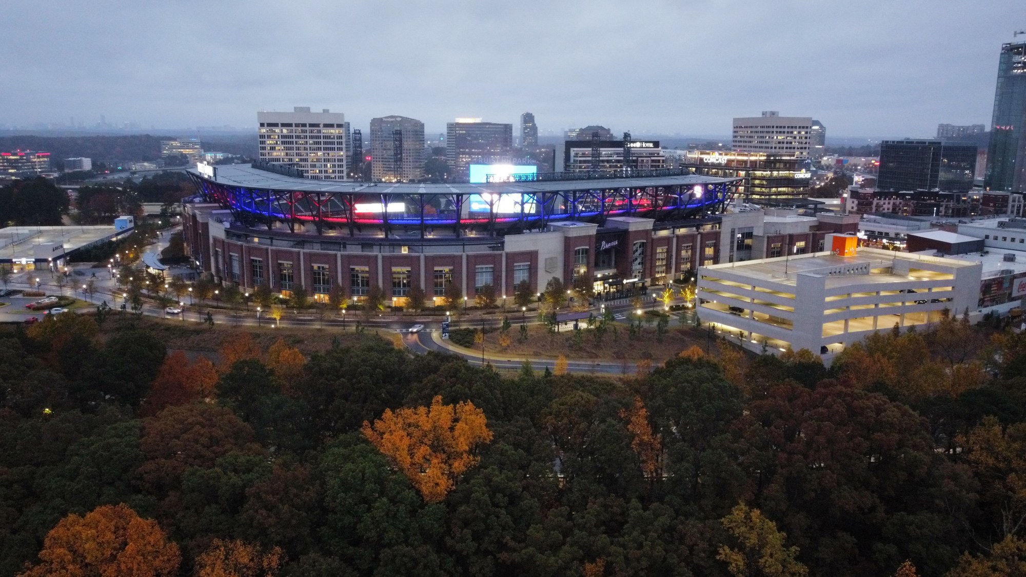 Aerial view at dusk of Truist Park, the Atlanta Braves' ballpark, lit in blue and red above autumn trees.