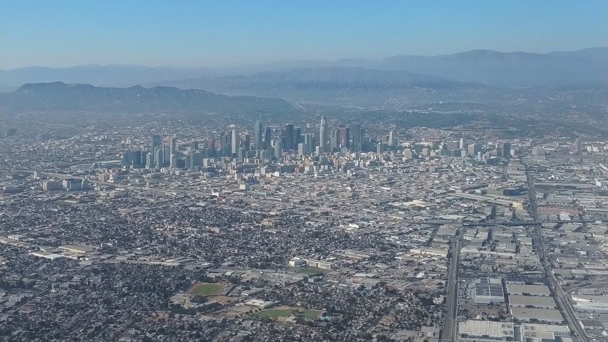 An aerial daytime view of downtown Los Angeles and surrounding neighborhoods under a hazy blue sky, with mountains in the distance.
