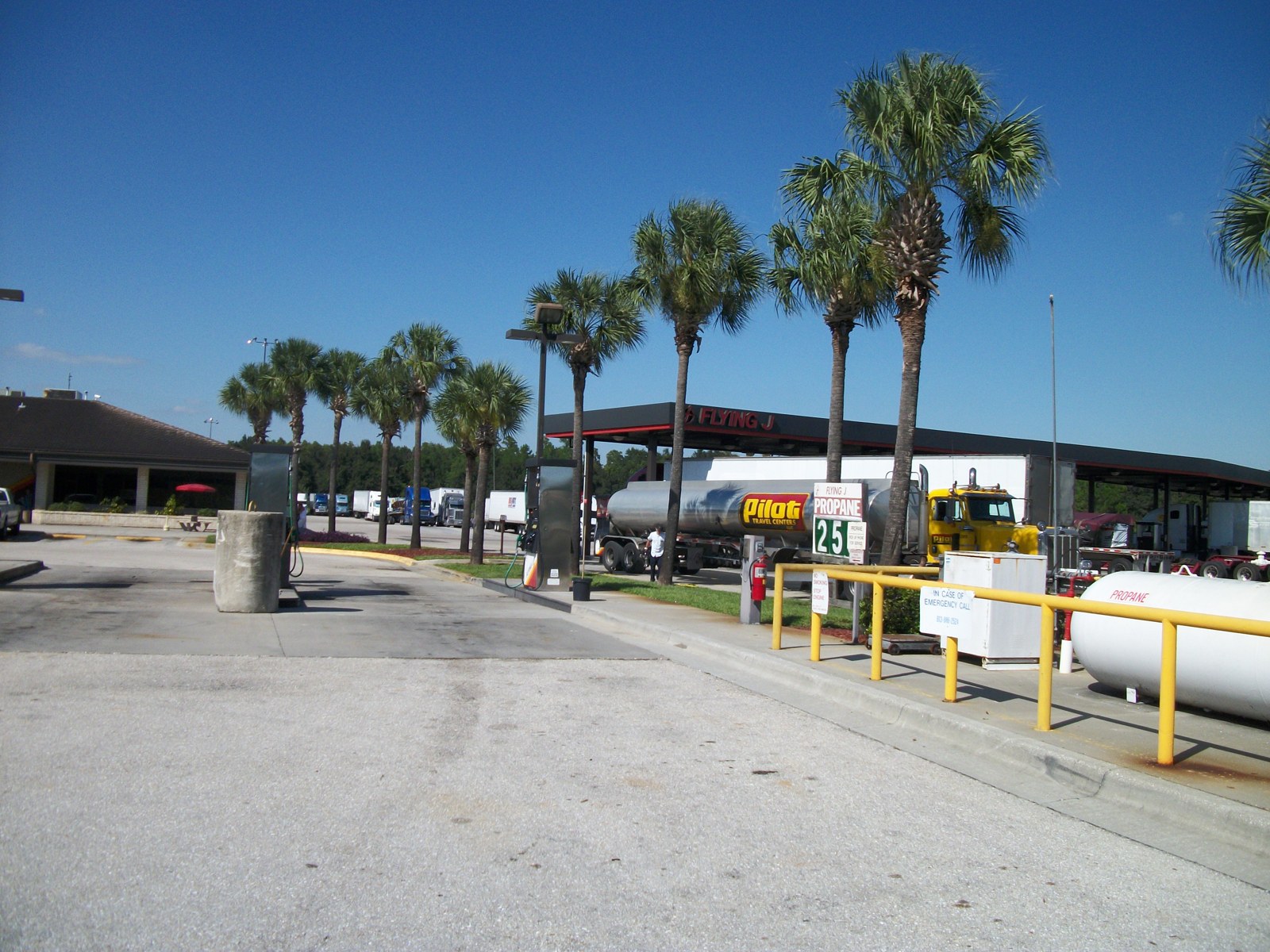A truck-stop fueling area under a blue sky, with a row of palm trees, a fuel pump, a propane tank and a silver tanker truck with a yellow cab parked beside the canopy.