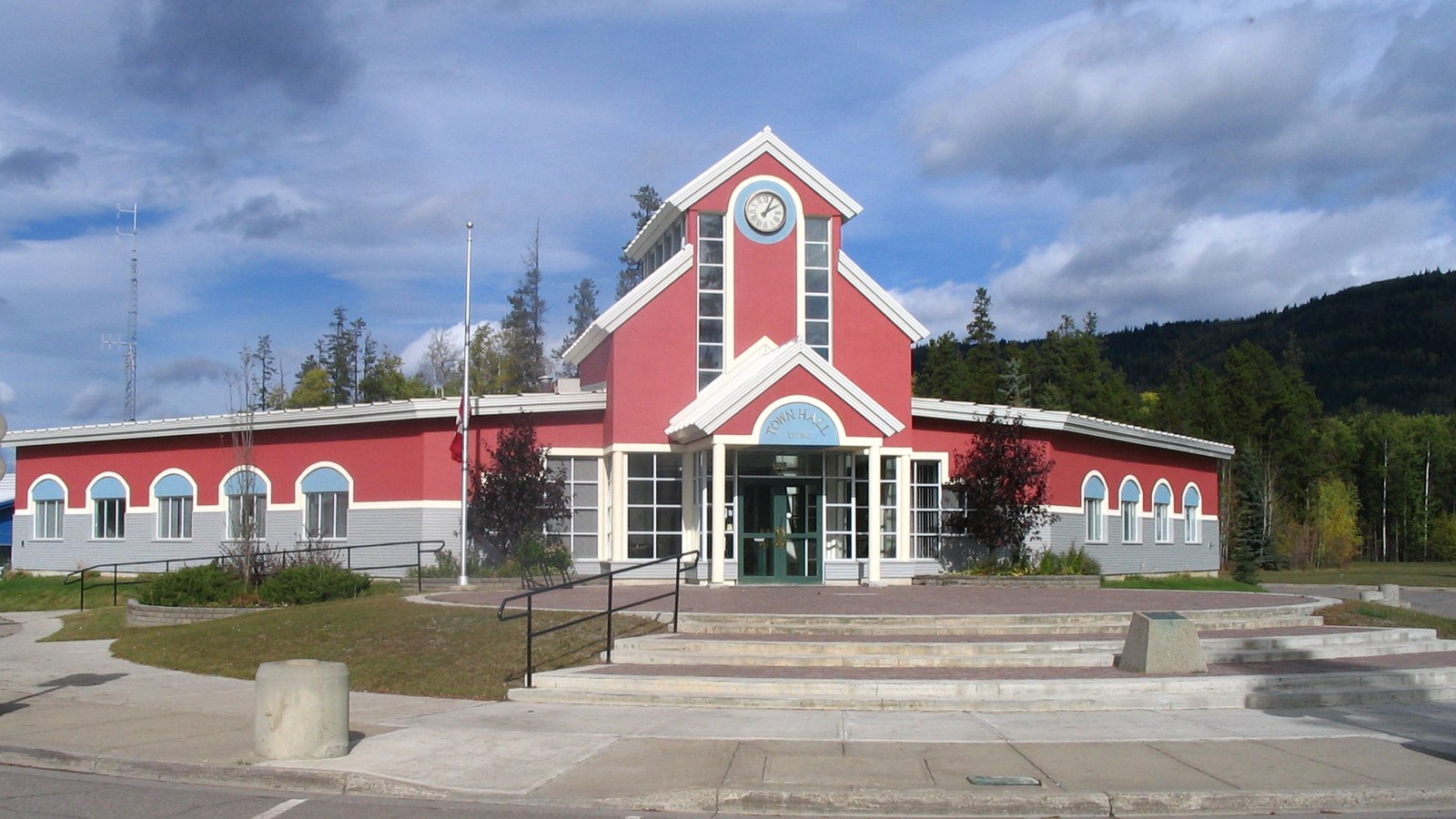 The red-and-white town hall of Tumbler Ridge, British Columbia, a one-storey building with a clock tower over its entrance, seen from the street under a cloudy sky with forested hills behind.
