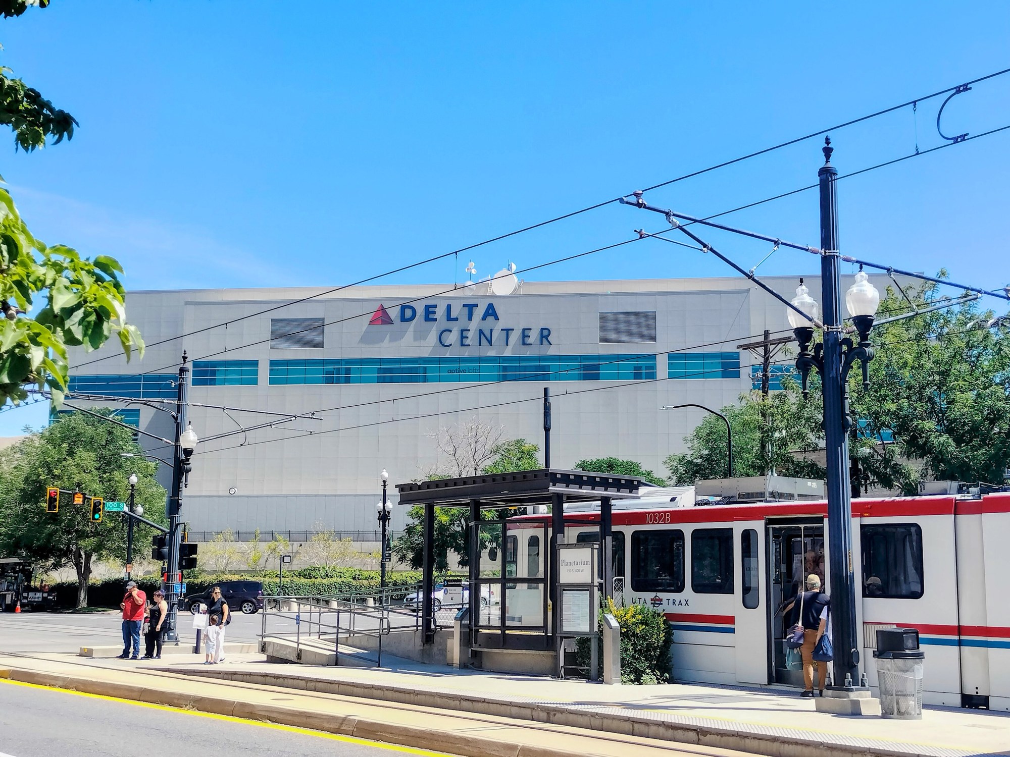The Delta Center arena in Salt Lake City on a sunny day, with a light-rail train and pedestrians in the foreground.