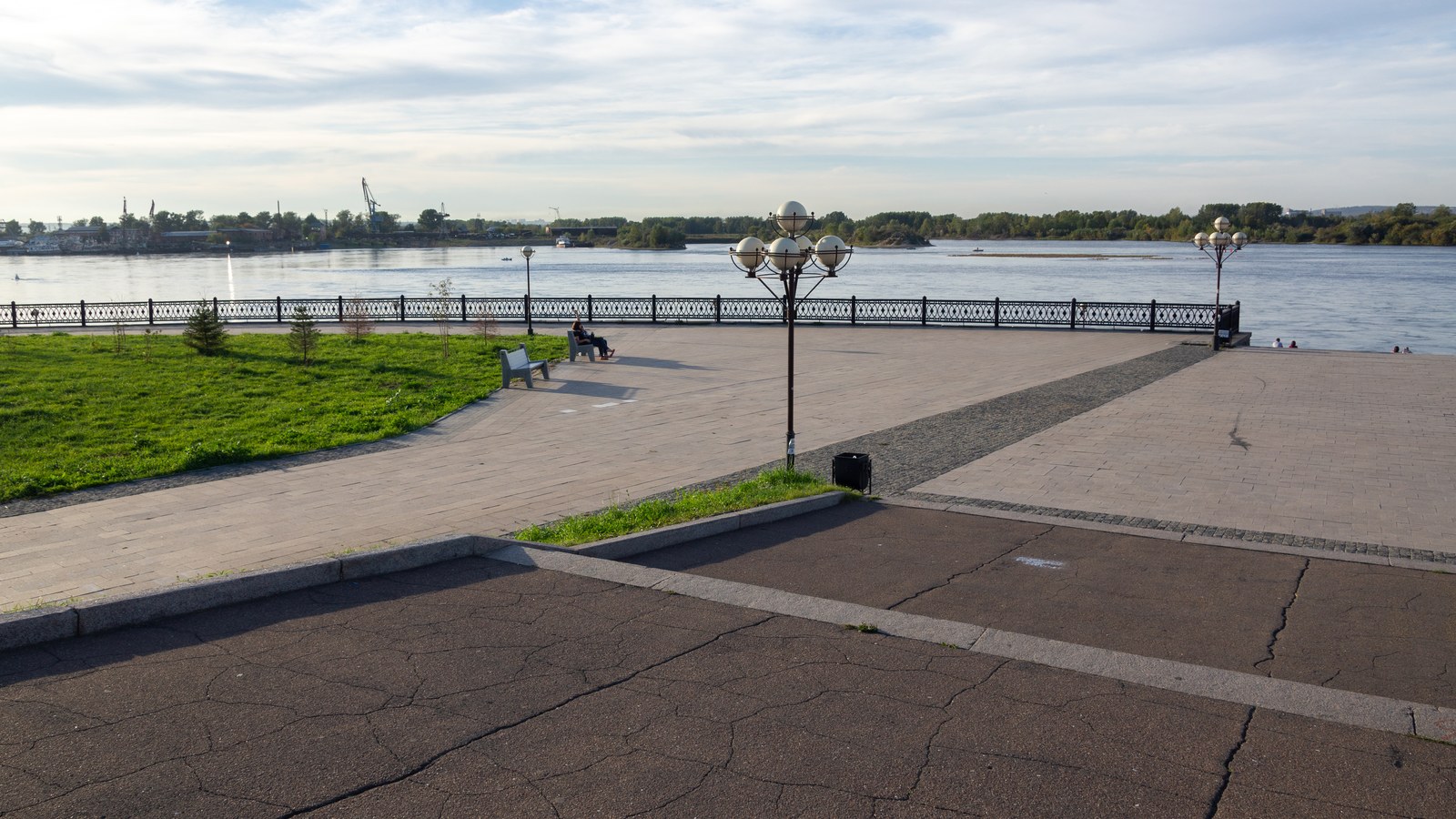 The Angara River embankment in Irkutsk, Russia, with a paved promenade, a lamp post with globe lights, benches and a wide river under a partly cloudy sky.