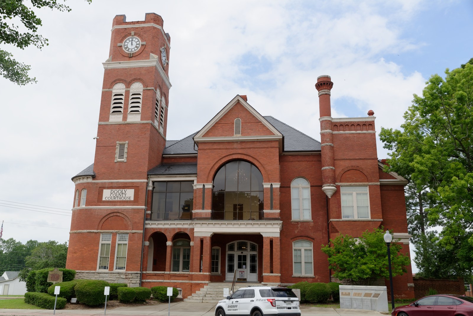 A red-brick county courthouse with a tall clock tower, a large arched window over the entrance and a white Dooly County Sheriff patrol vehicle parked in front.