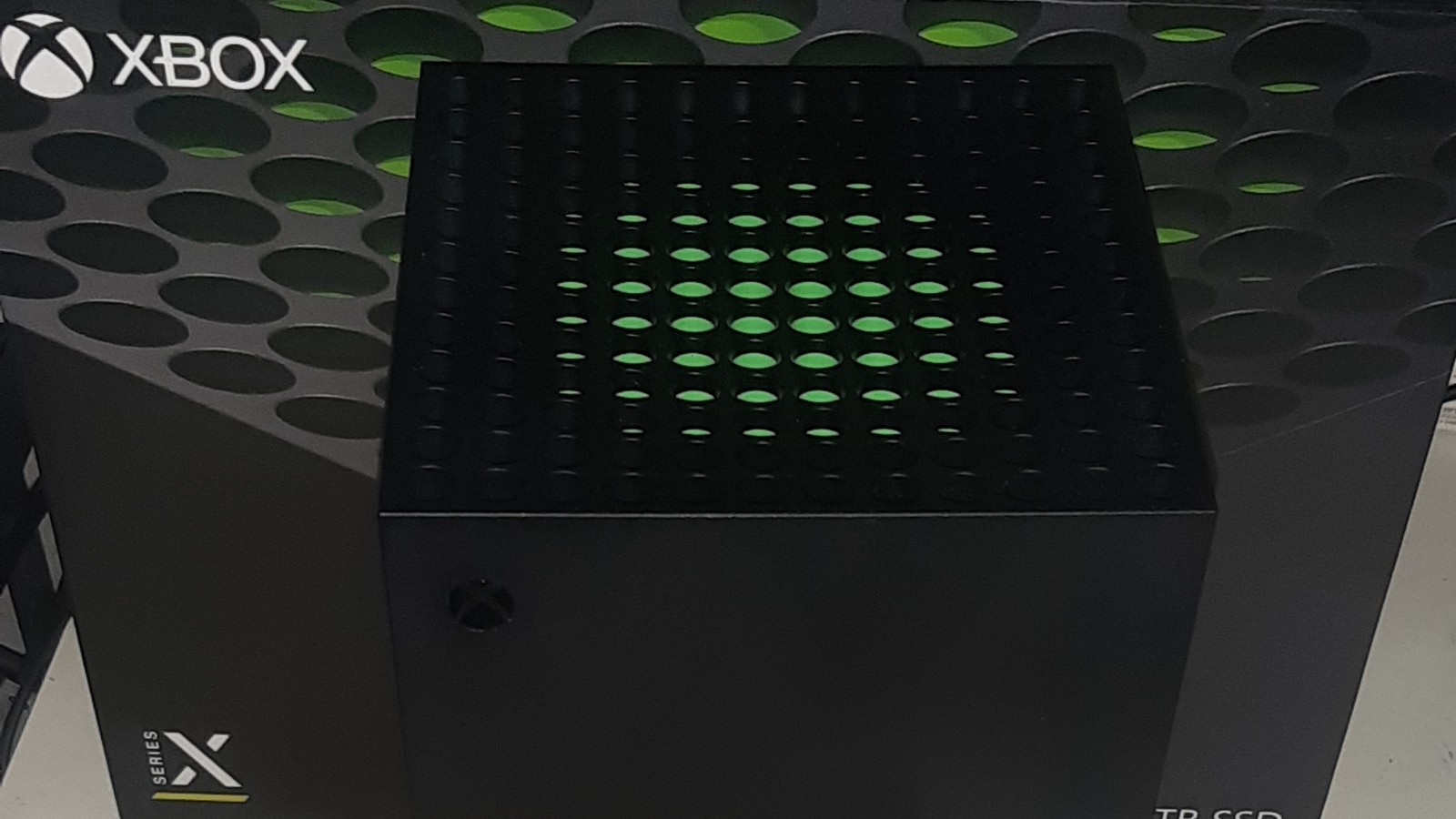 A close view of the top of an Xbox Series X console, a black tower with green light showing through its vents, in front of a retail box with the Xbox logo.