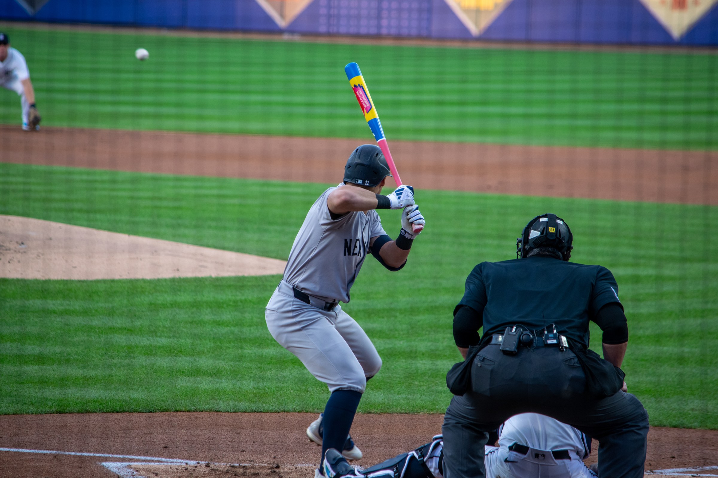 A New York Yankees batter, seen from behind, in a batting stance with a colorful bat while the catcher and umpire crouch behind the plate.
